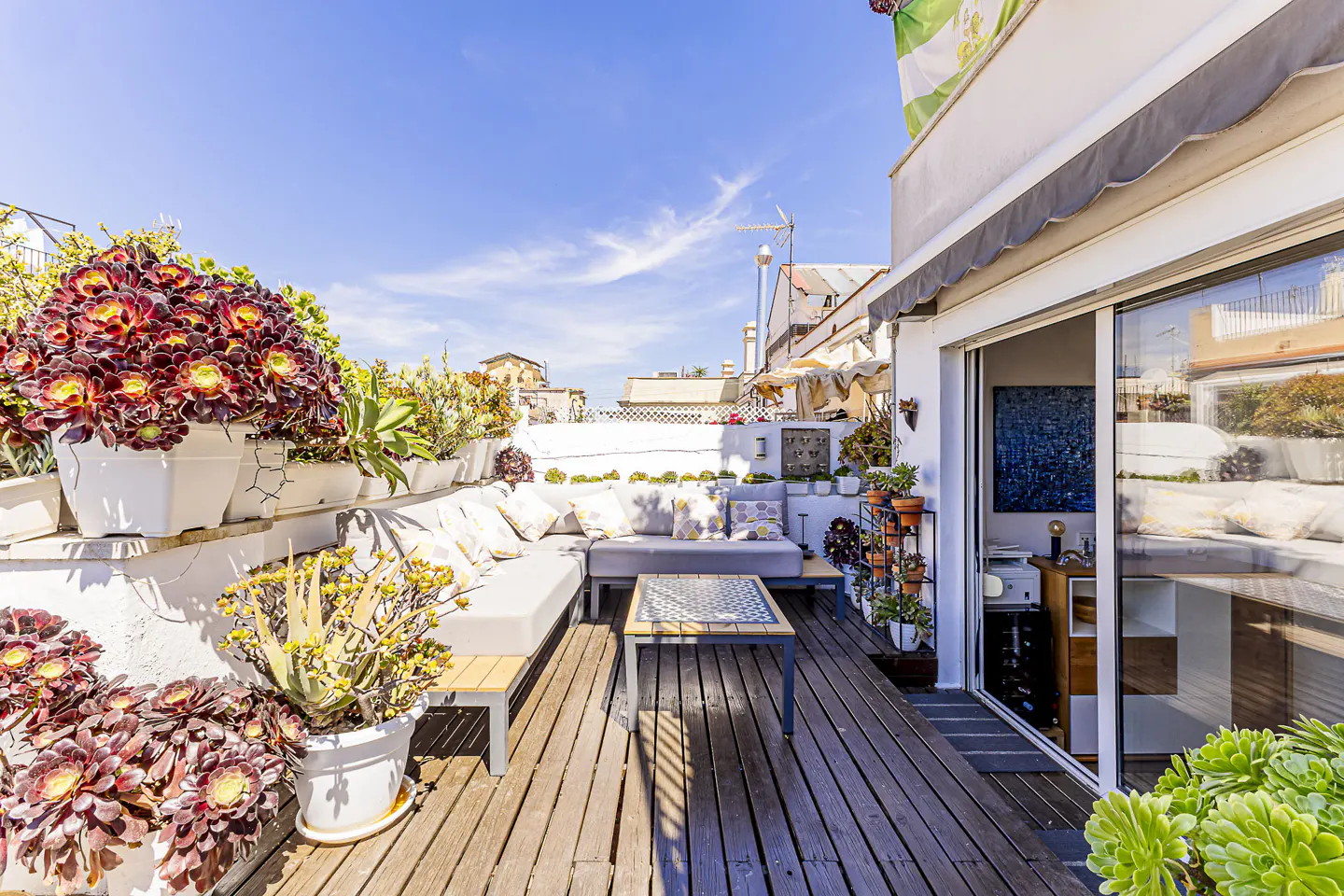 Sunny rooftop patio with wooden deck, gray sectional sofa, and potted plants. Sliding glass doors lead inside. Blue sky above.