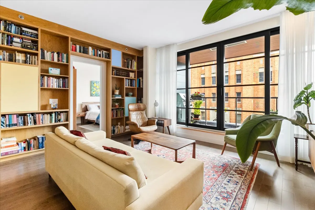 Bright living room with built-in bookshelves, a cream sofa, and a large window with a view of a brick building. A doorway leads to a bedroom.