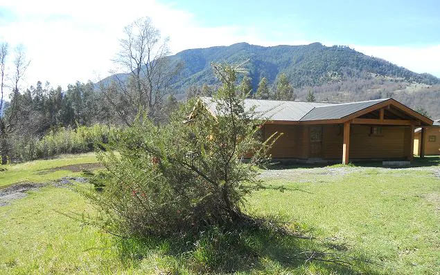 A wooden cabin with a gray roof sits on a green lawn, with a mountain backdrop under a blue sky.