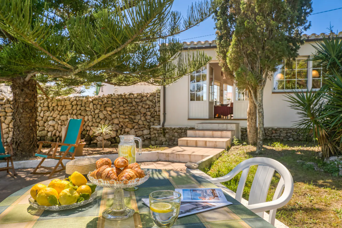 Outdoor dining scene: Table with croissants, lemons, and lemonade. Blue chairs and a stone wall are in the background.