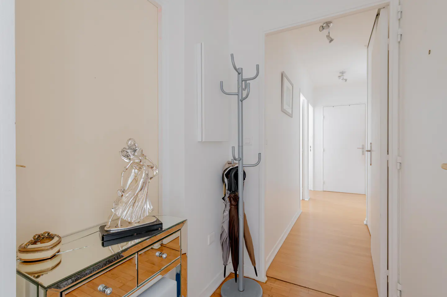 A mirrored console table with a silver sculpture stands in a hallway next to a coat rack and a view into a bright corridor.