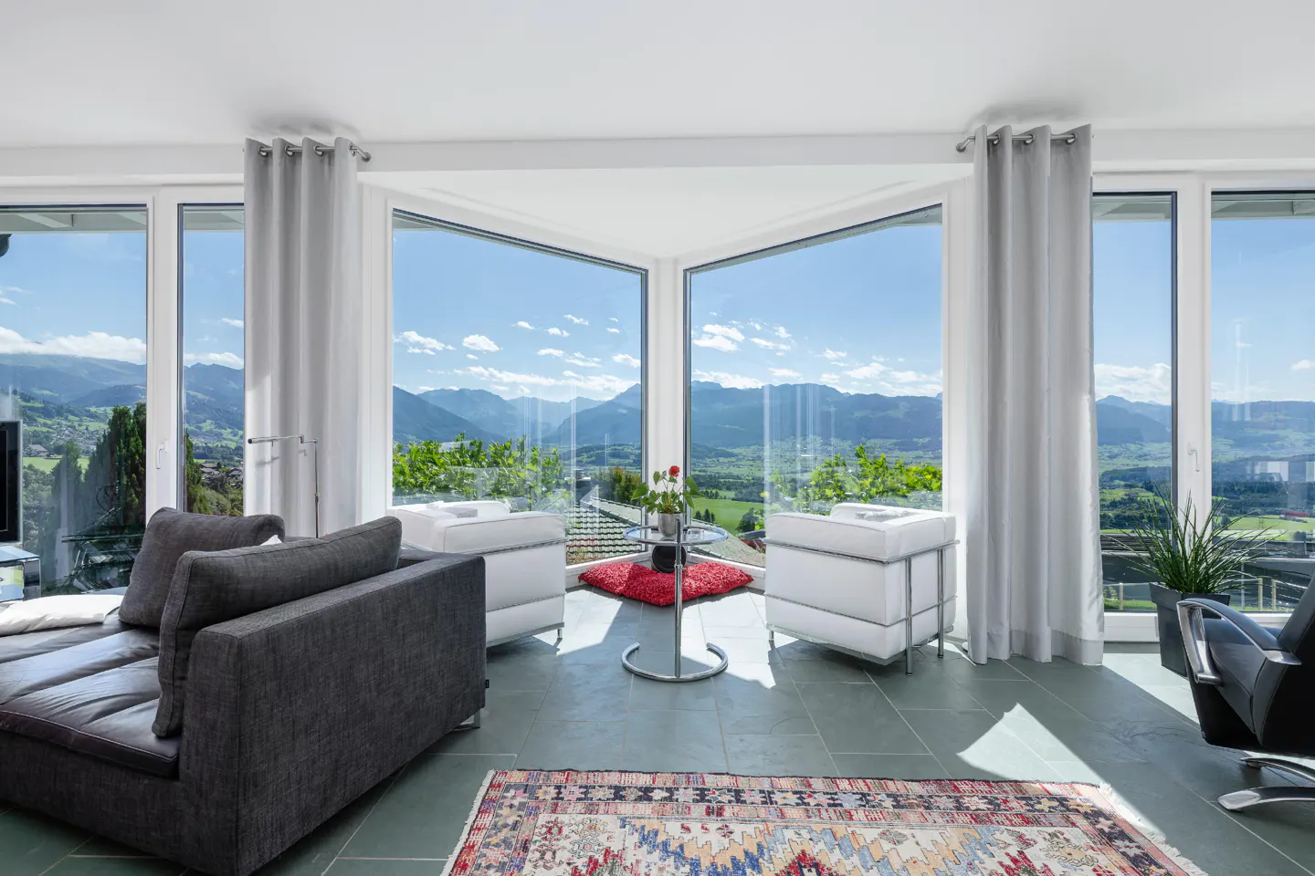 Living room with gray sofa, white chairs, and large windows showcasing a mountain view. A colorful rug adds warmth to the gray tile floor.