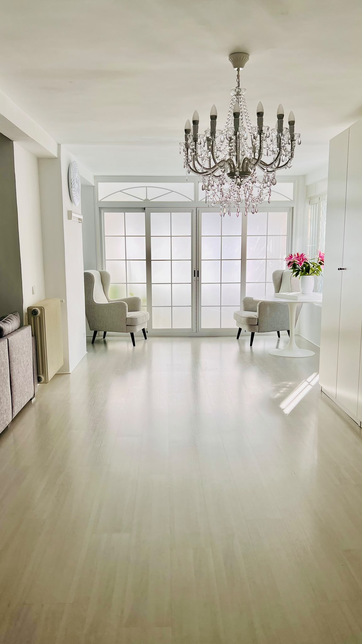 Bright, minimalist living room with light wood floors, a crystal chandelier, two armchairs, and large glass patio doors. [AI-generated text]