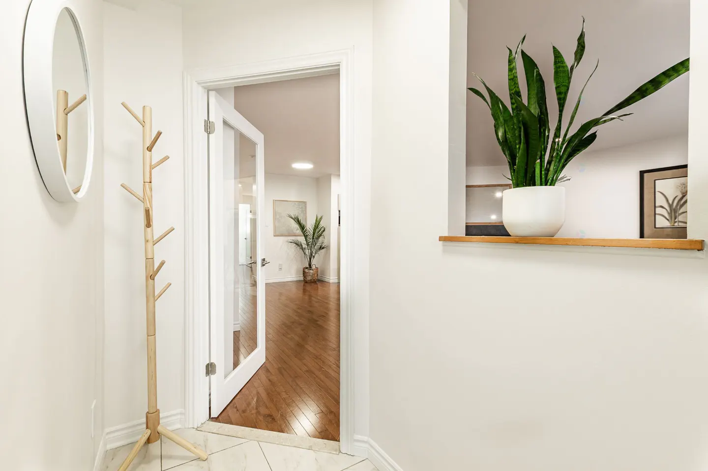 Bright entryway with white walls, marble tile, and wood floors. A coat rack and mirror are on the left, and a snake plant sits in a niche on the right.