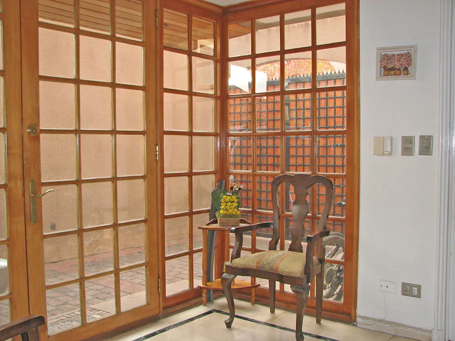 A sunroom with wooden framed windows, a wooden chair with a patterned cushion, and a small table with yellow flowers.