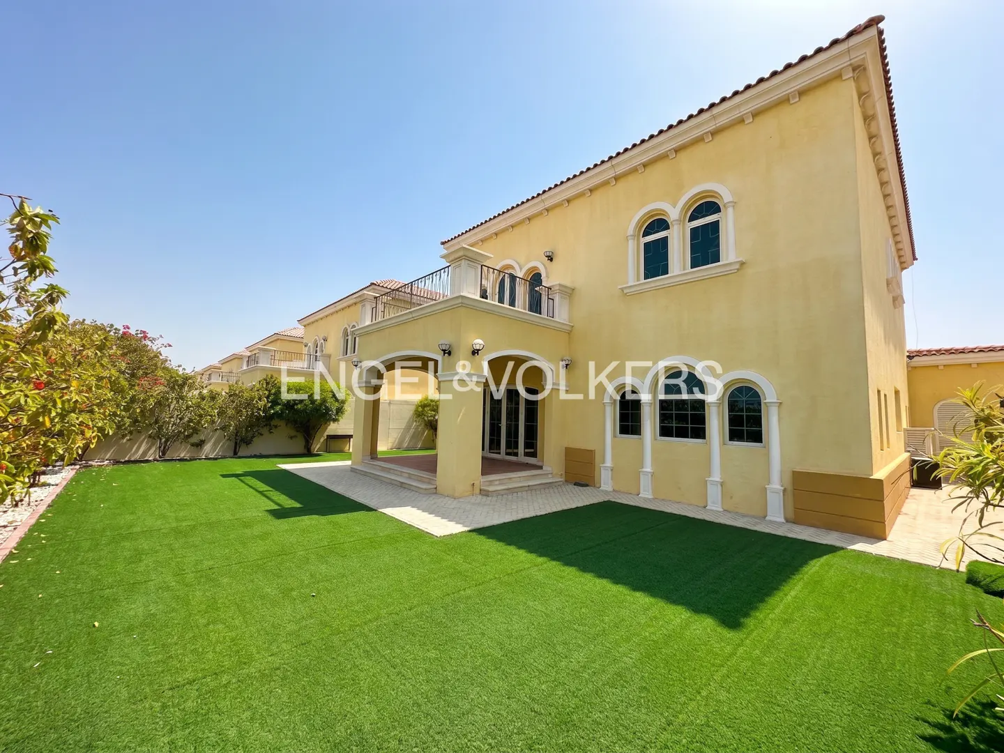 A two-story yellow villa with arched windows, a balcony, and a covered patio overlooking a large green lawn.