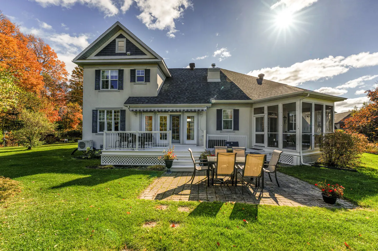 Backyard view of a two-story house with a patio set on a brick surface and a screened-in porch. The lawn is green, and trees are in the background.