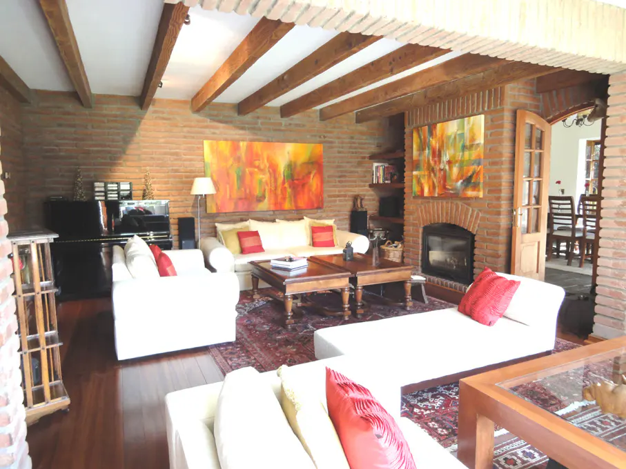 A living room with brick walls, wooden beams, white sofas, a fireplace, and colorful paintings.
