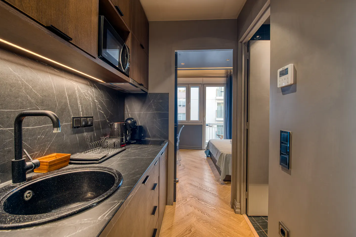 A modern apartment features a black marble kitchen with a sink, and a view into a bedroom with a balcony.