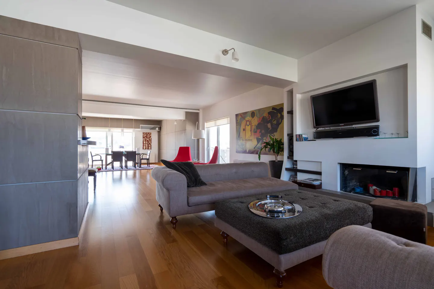 Open-concept living room with hardwood floors, gray sofa, ottoman, and fireplace with TV above. Dining area visible in background.