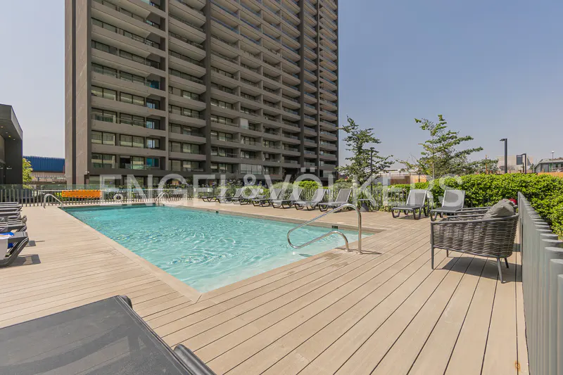 Outdoor pool with lounge chairs on a wooden deck, next to a tall apartment building on a sunny day.