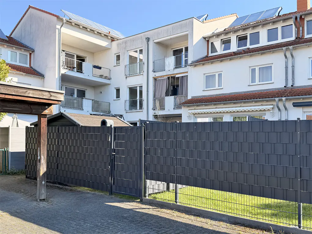 Exterior view of a modern, multi-story apartment building with balconies and a gray privacy fence in front.