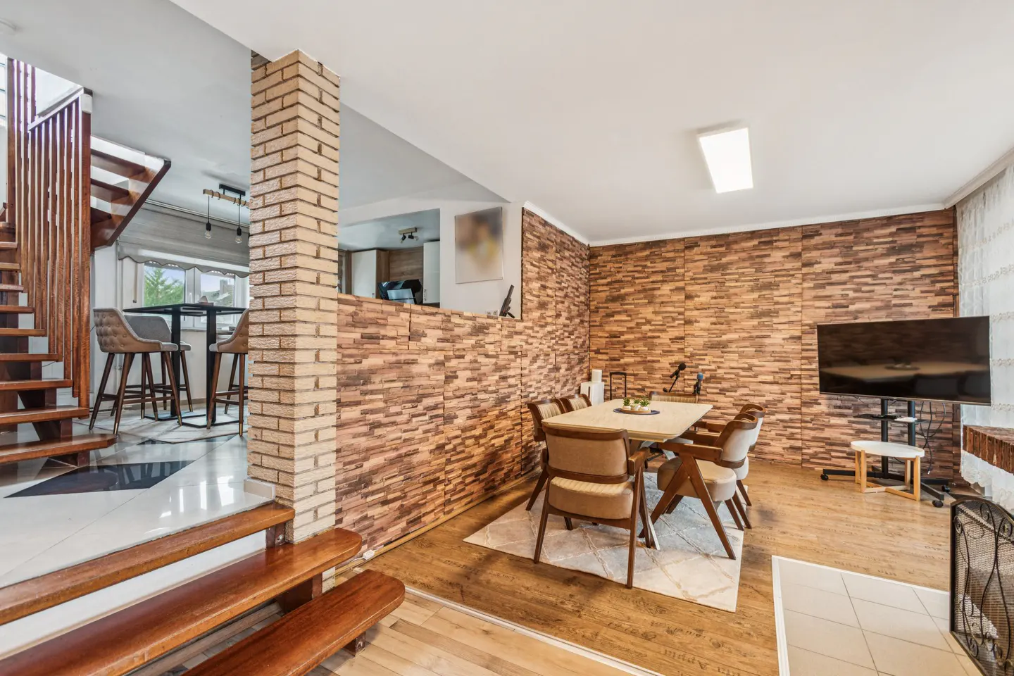 Open-concept living space with wood floors, brick accent wall, dining table, and stairs leading to an upper level.
