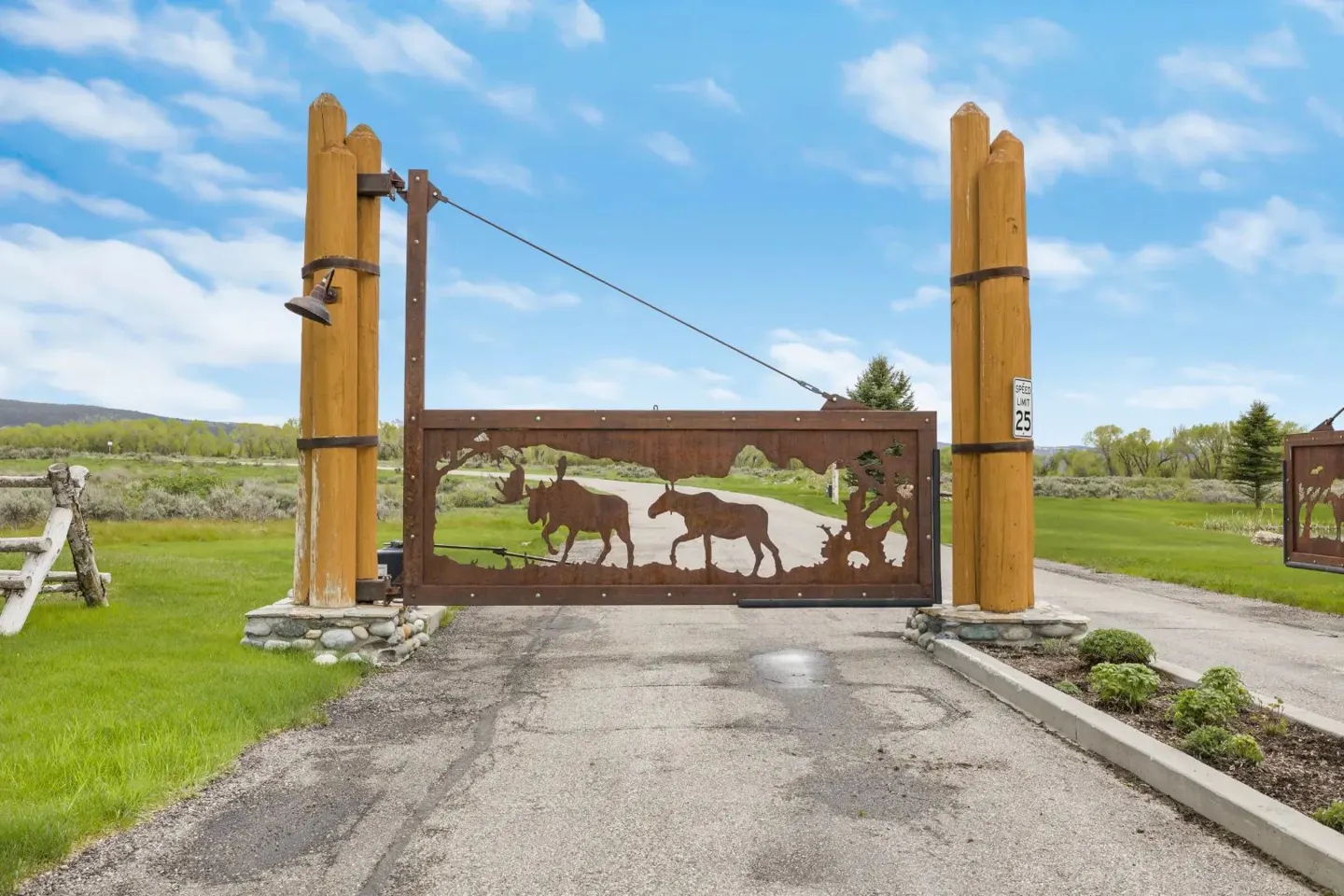 A metal gate featuring moose silhouettes between two tall wooden posts at the entrance of a rural property.