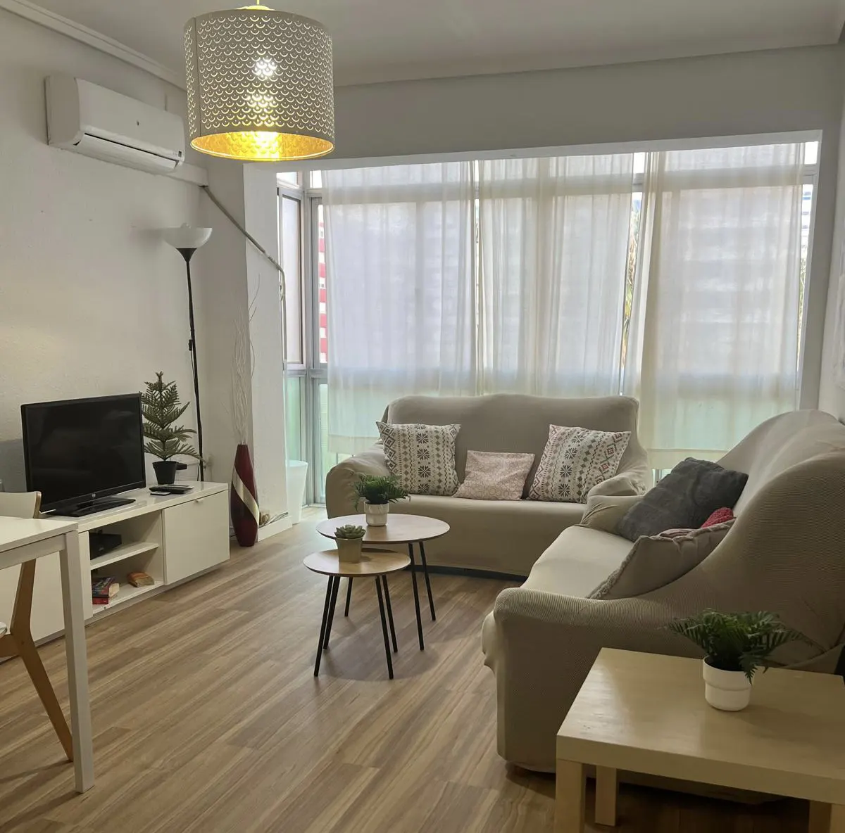 Bright living room with light wood floors, two beige sofas, nested coffee tables, a TV console, and sheer window curtains.