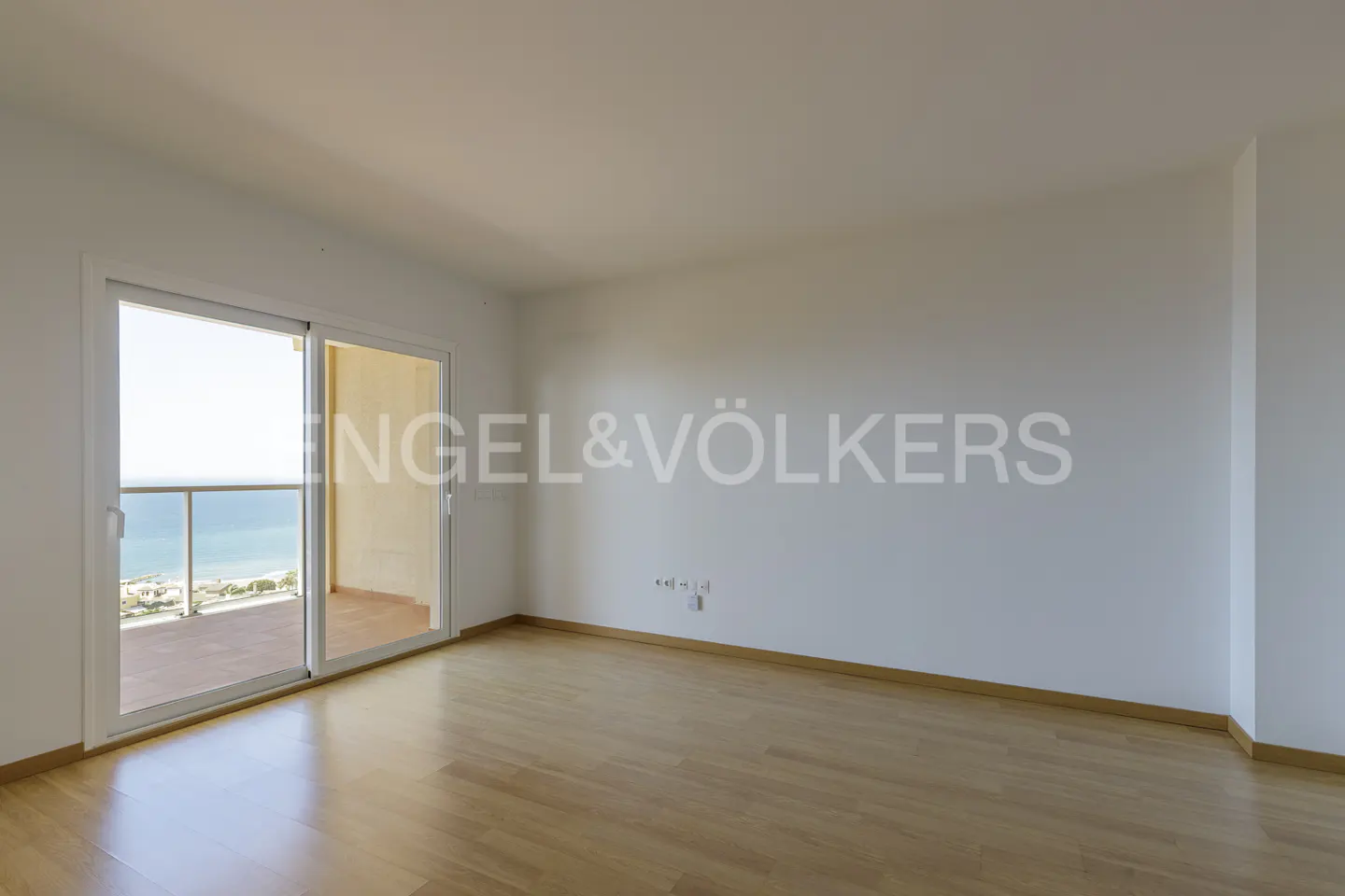 Empty room with light wood floors, white walls, and sliding glass doors opening to a balcony with a view of the ocean.