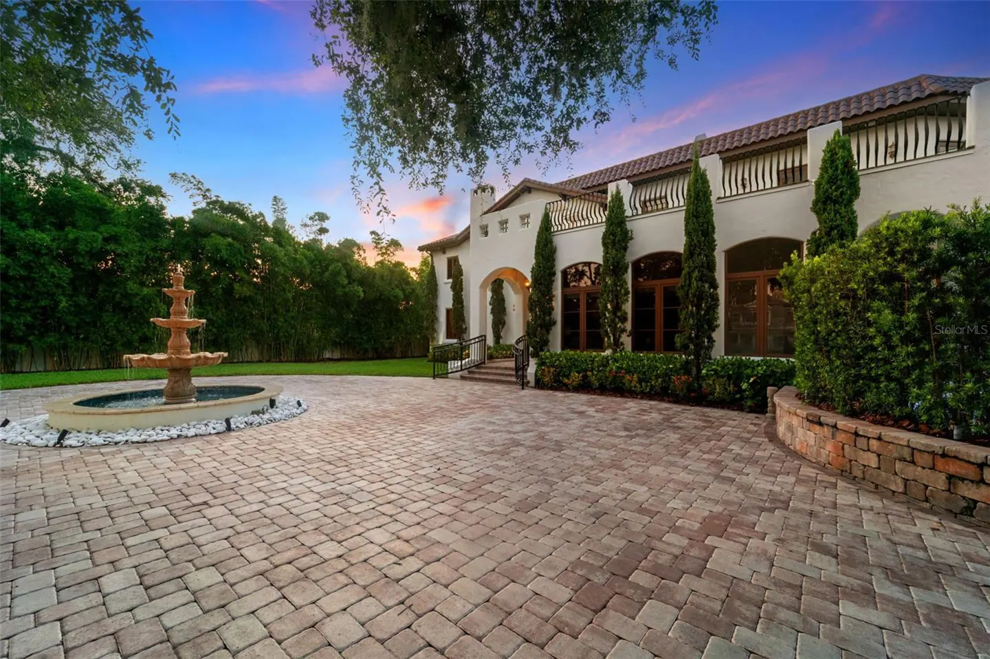 Exterior of a large, white house with a red tile roof and a brick driveway. A fountain is on the left.
