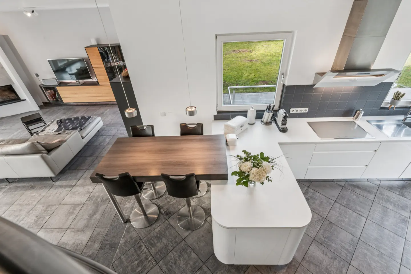 Open-concept kitchen and living room with a wood table, white cabinets, and gray tile flooring. A white sofa is visible in the background.