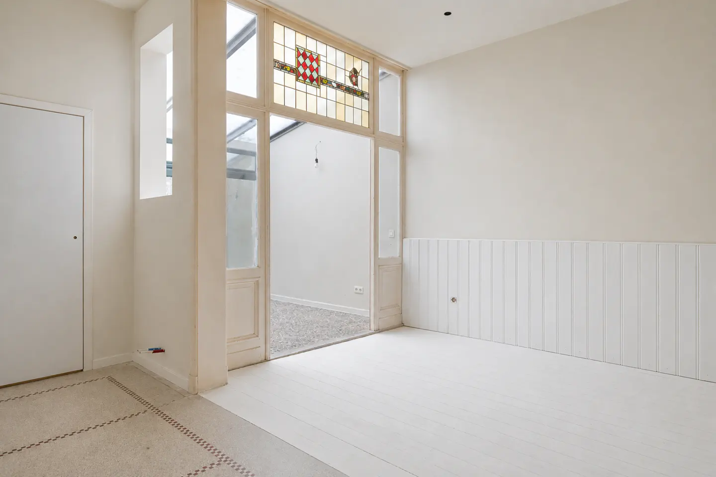 Bright, empty room with white walls, floors, and wainscoting. A doorway with stained glass leads to another room with gray carpet.