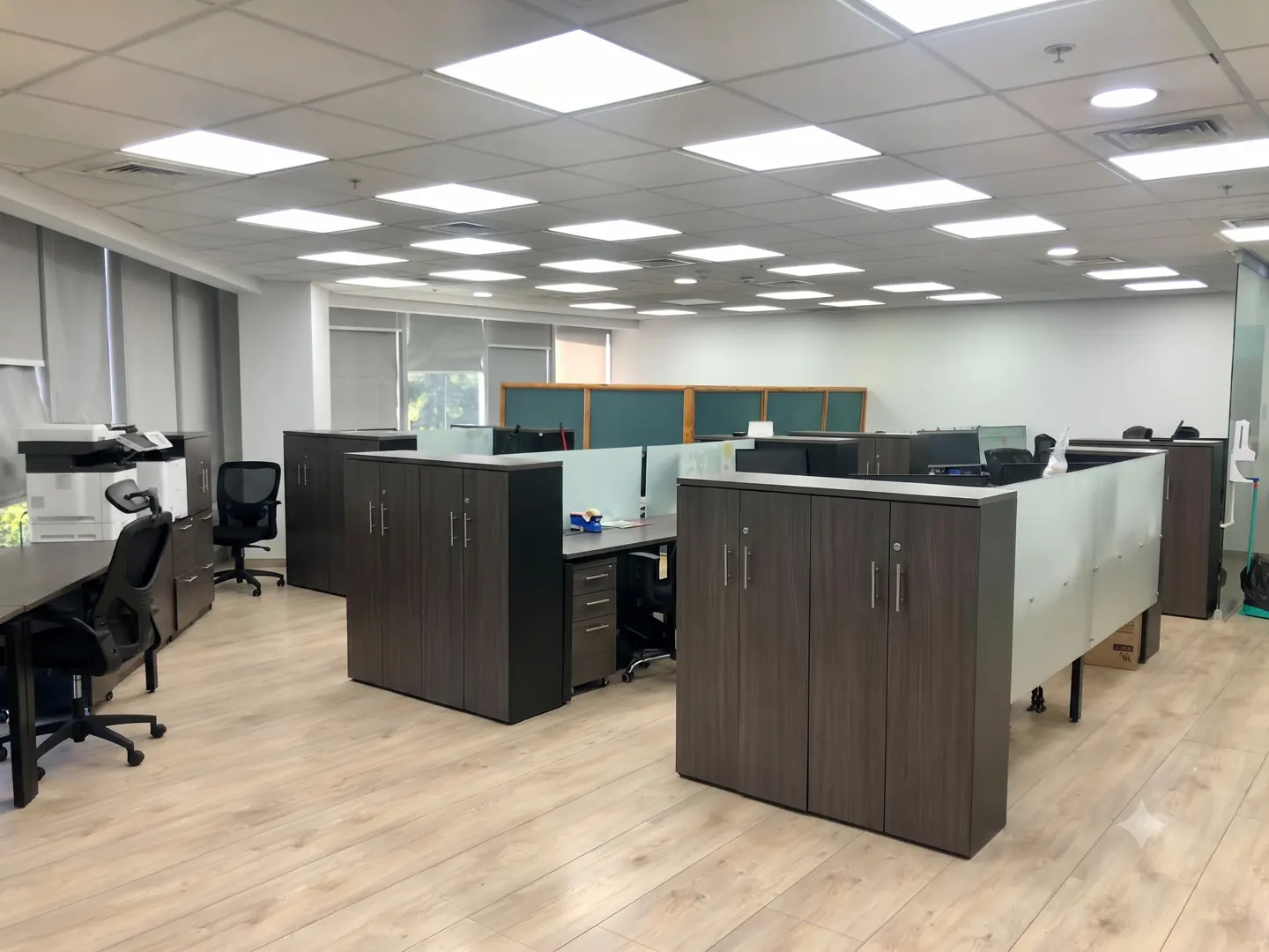 Modern office space with dark wood cubicles, light wood flooring, and bright recessed ceiling lights.
