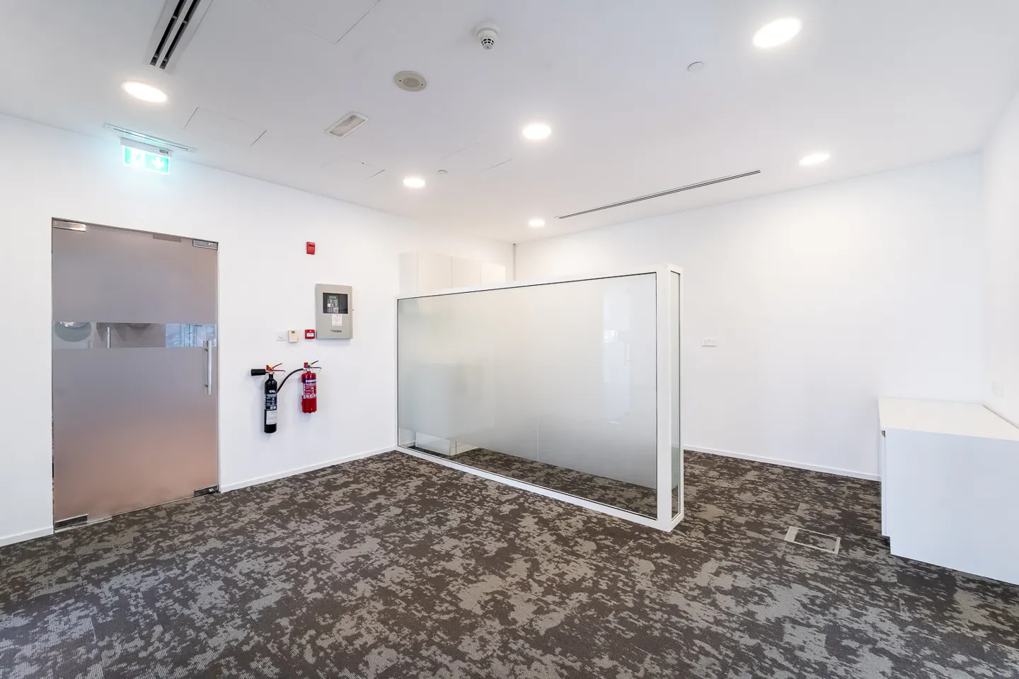 Bright, empty office space with white walls, patterned carpet, and a frosted glass partition. A glass door and fire extinguishers are visible.