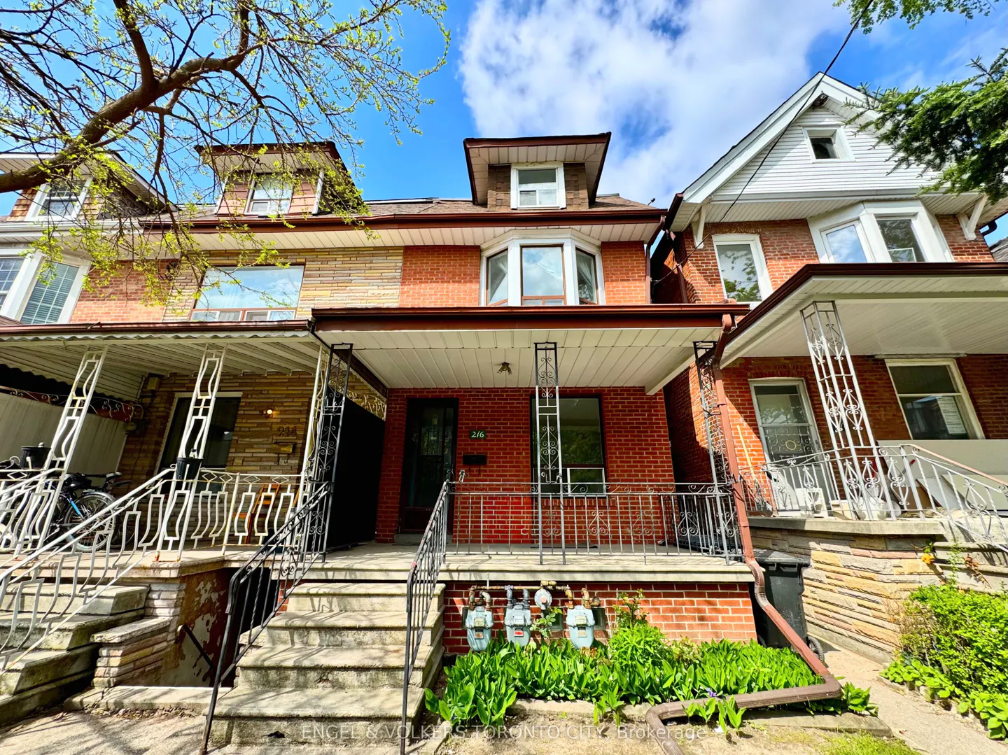 A three-story red brick house with a porch and black railings, set against a blue sky with clouds.