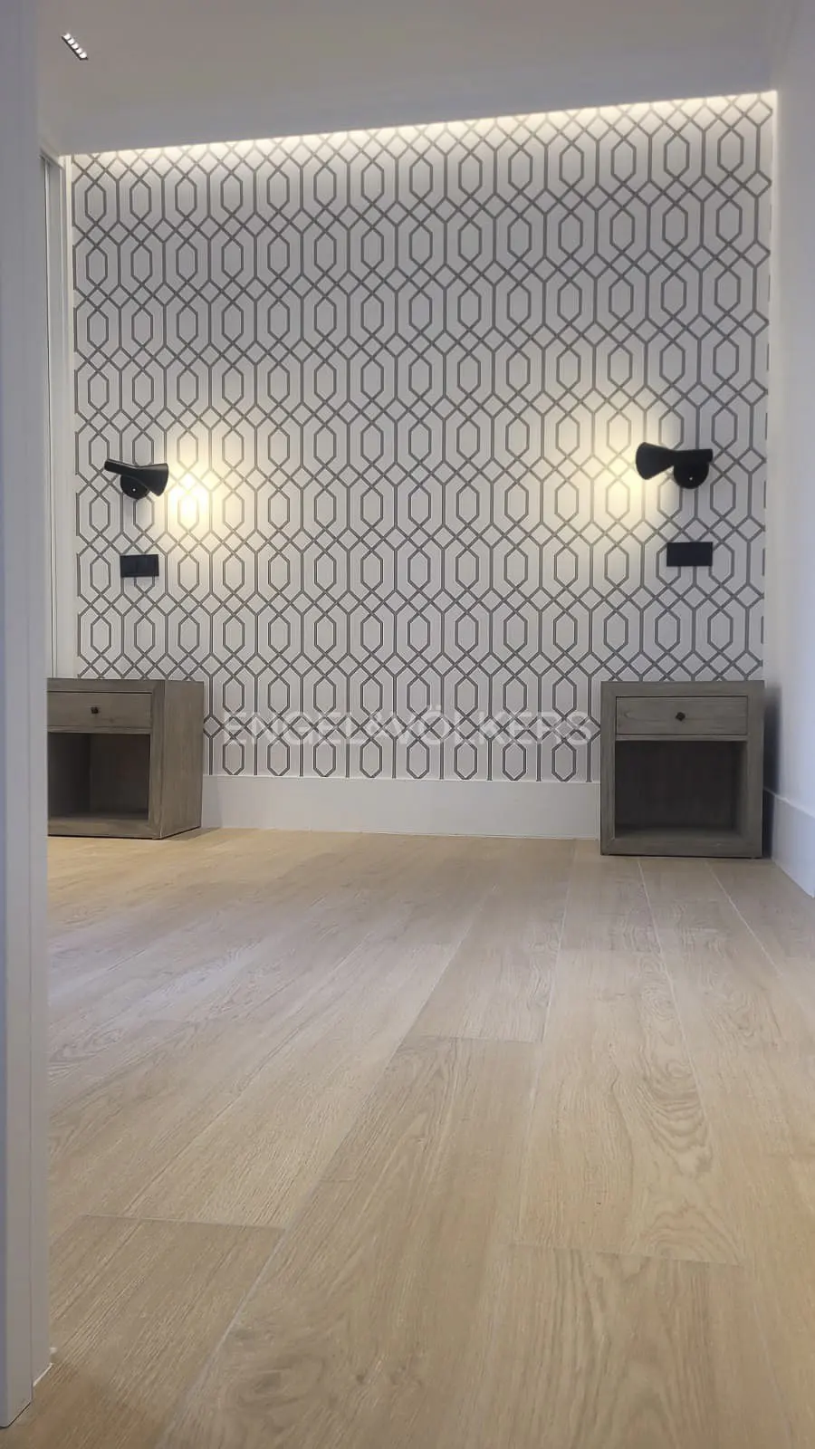 Bedroom with light wood floors, geometric wallpaper, and matching nightstands with black sconces.