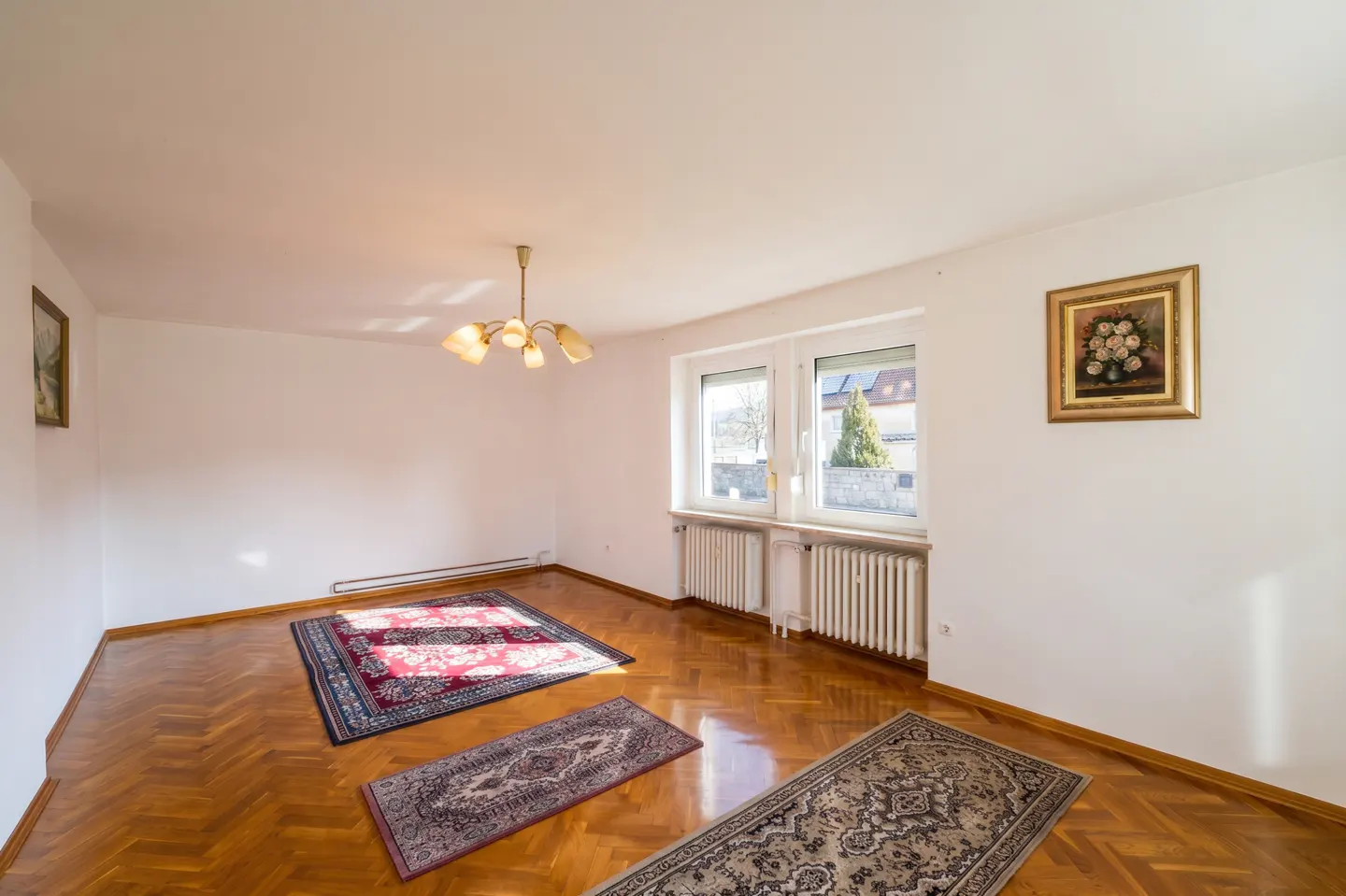 Empty room with white walls, herringbone wood floors, three patterned area rugs, two windows, and a vintage chandelier.