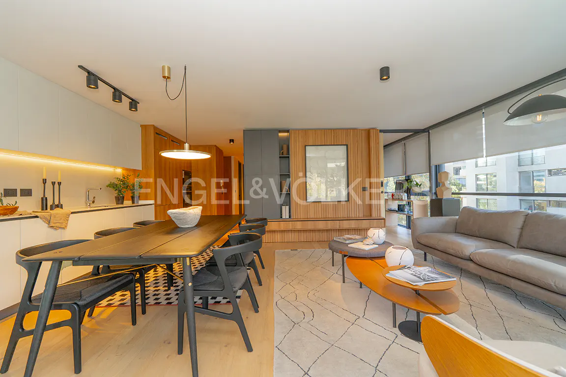 Open-concept living space with a black dining table, gray sofa, and light wood accents. Large windows offer natural light.