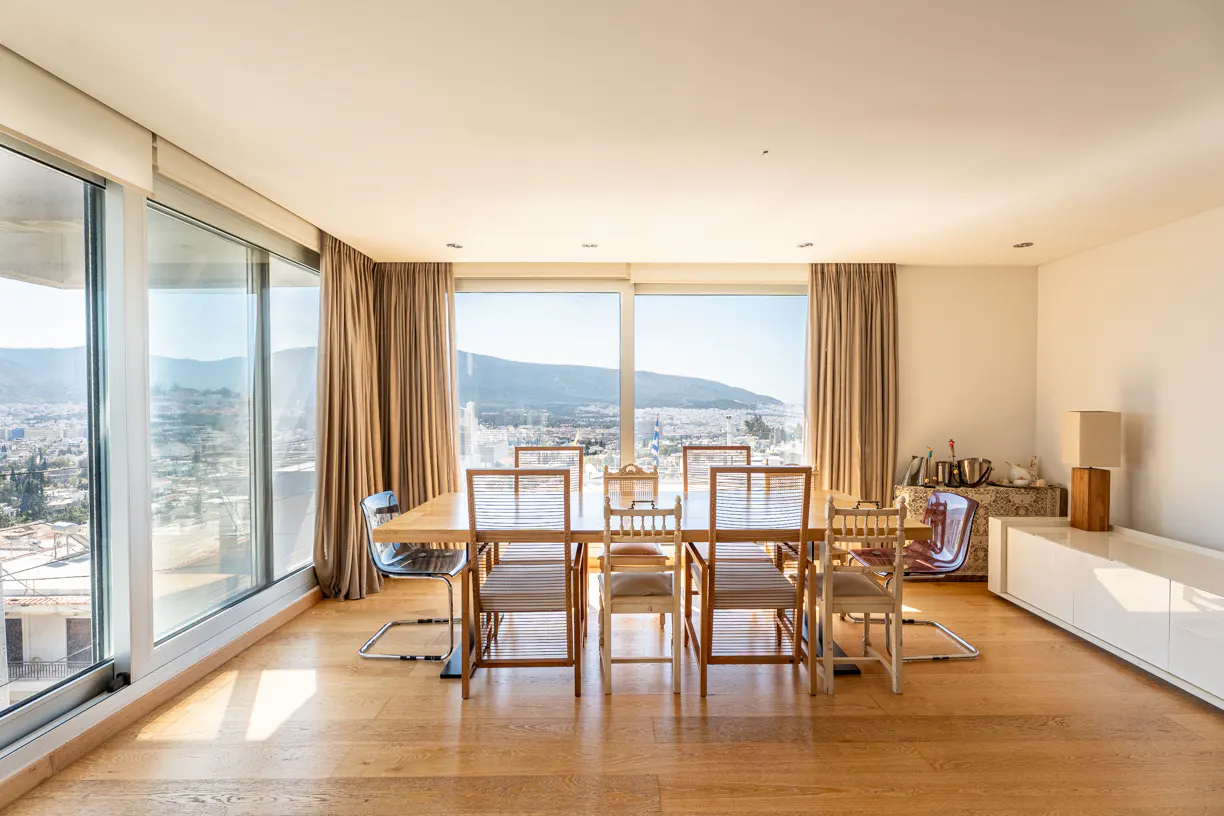 Bright dining room with wood floors, table, and chairs. Large windows offer a city view. Beige curtains frame the windows.