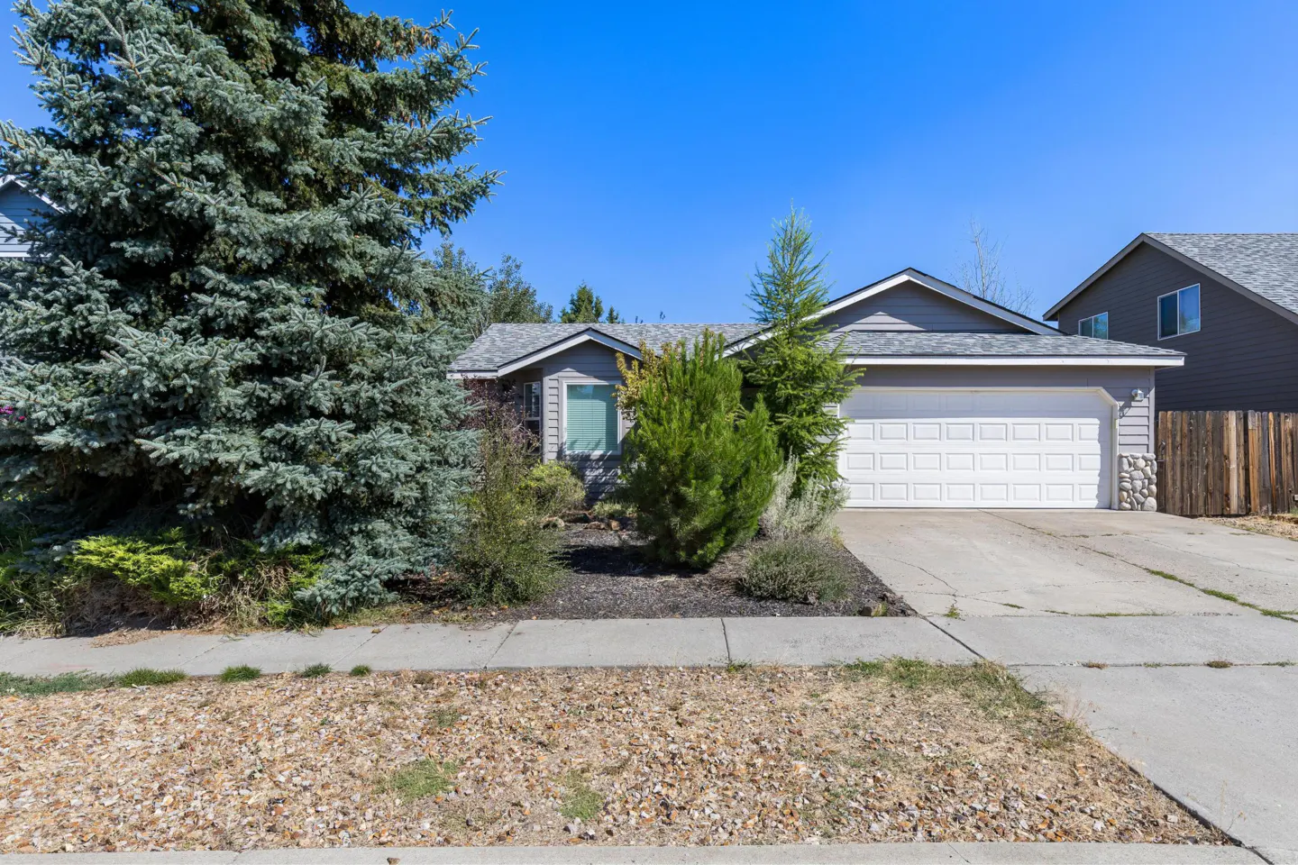 A single-story gray house with a white garage door, green trees, and a blue sky.