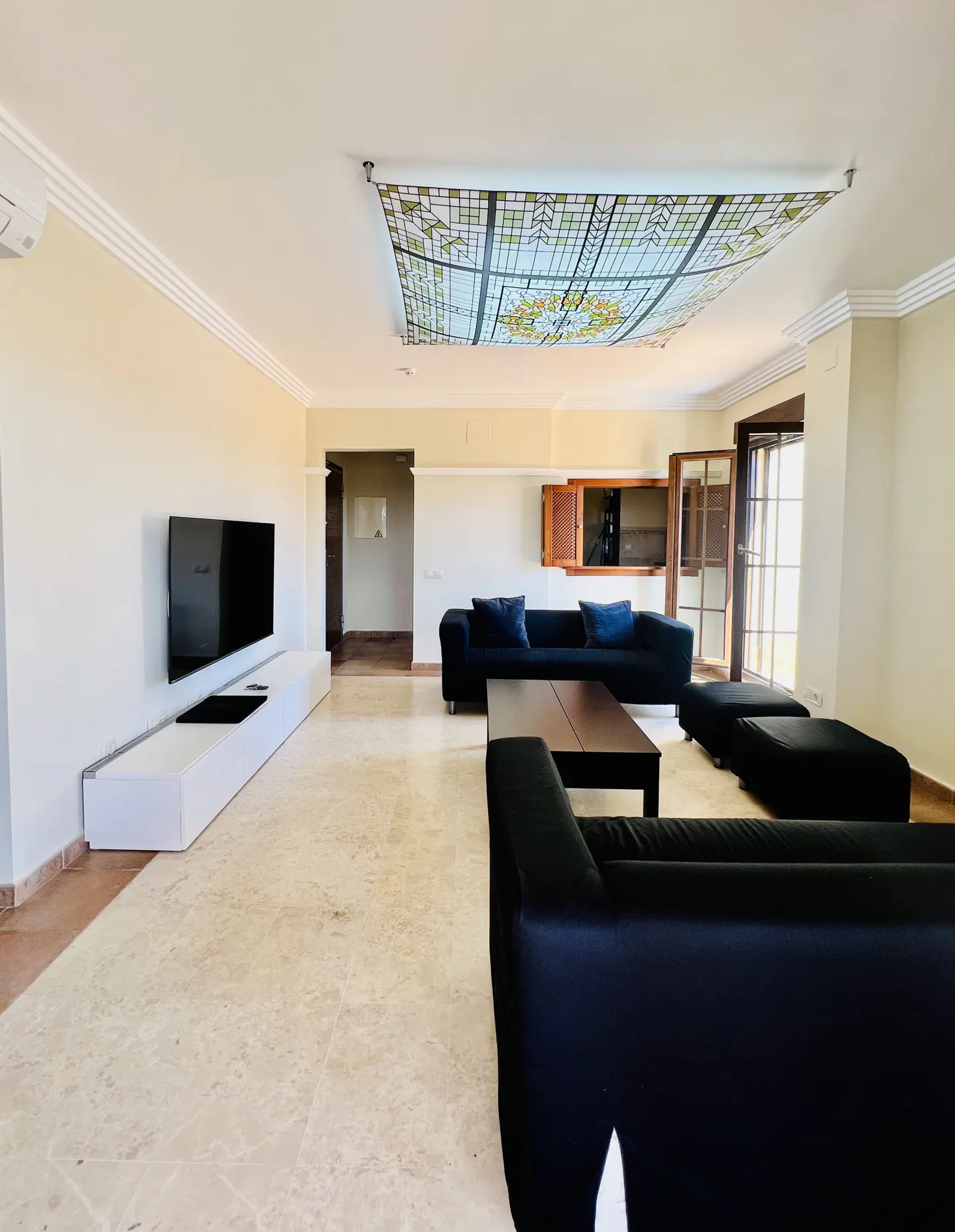 A spacious living room with black sofas, a dark wood coffee table, and a large, decorative stained-glass ceiling panel.