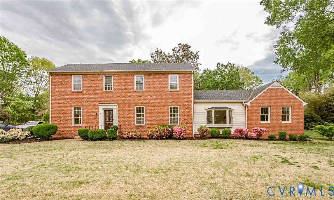 Two-story brick house with a dark roof, white trim, and a green lawn. Bushes and pink flowers line the front of the house.