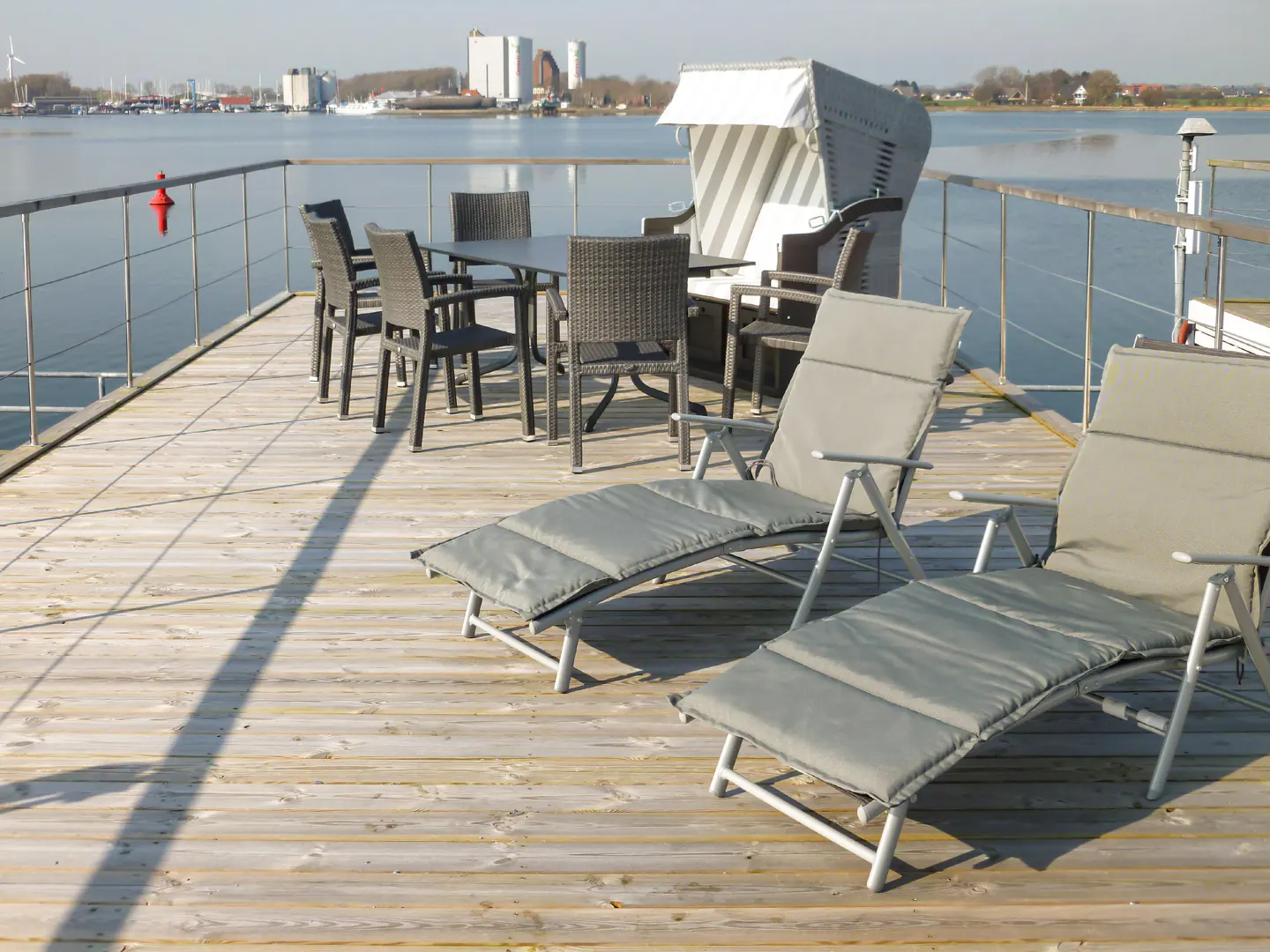 Outdoor patio on a wooden deck overlooking water. Two lounge chairs, a table with chairs, and a hooded beach chair are visible.