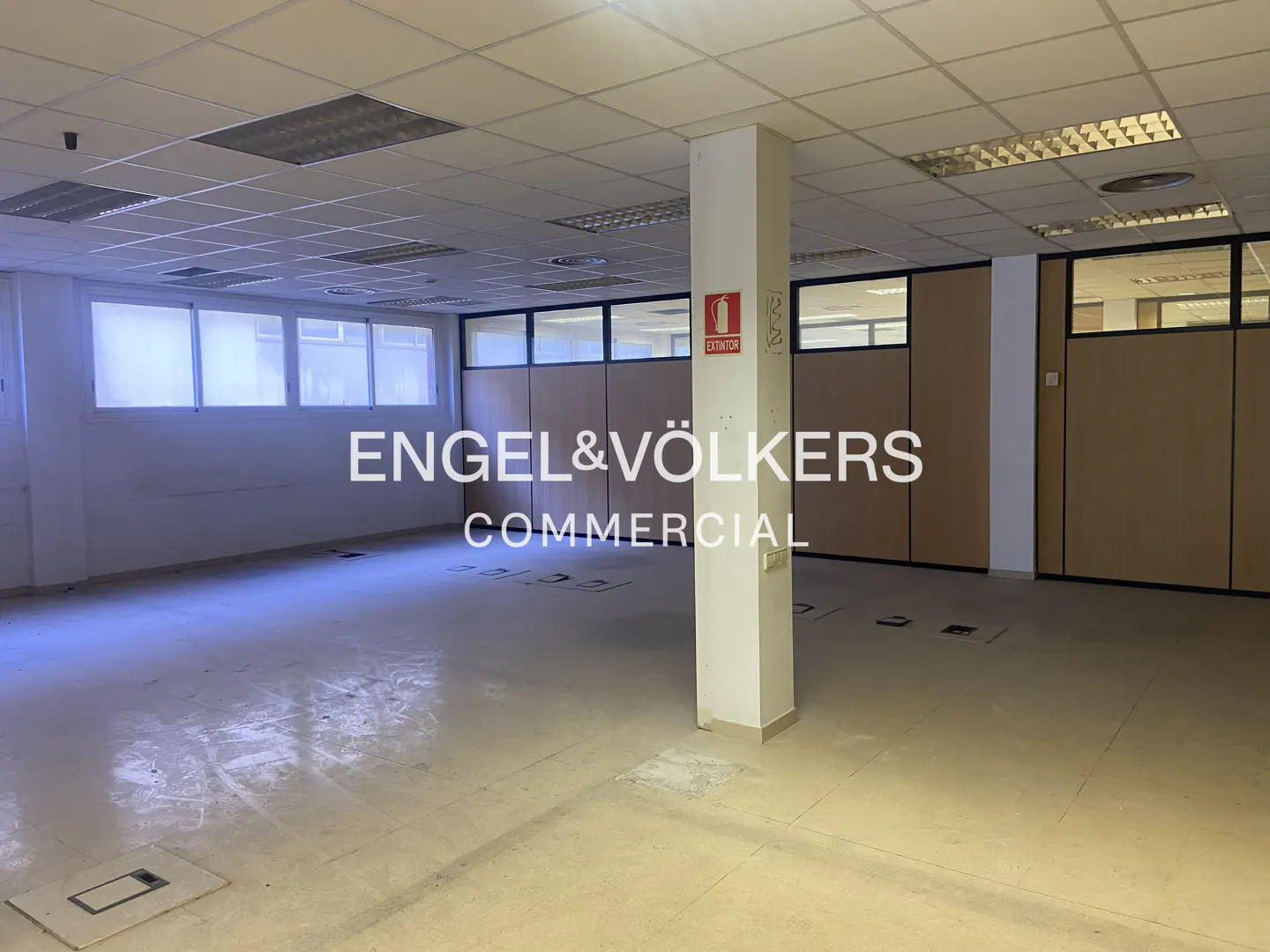 Empty office space with white walls, a tiled ceiling, and a support column with a fire extinguisher sign.