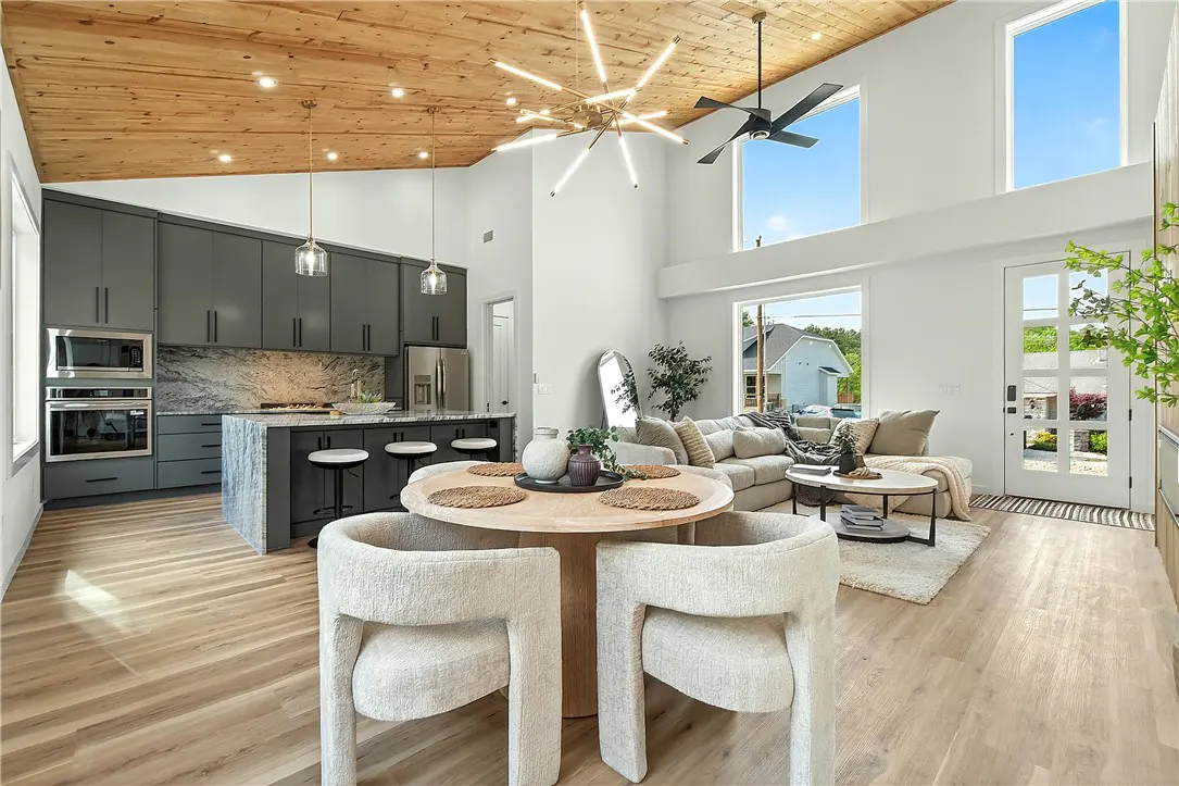 Open-concept living space with a kitchen, dining area, and living room. Gray cabinets, wood ceiling, and large windows. Modern lighting fixtures.