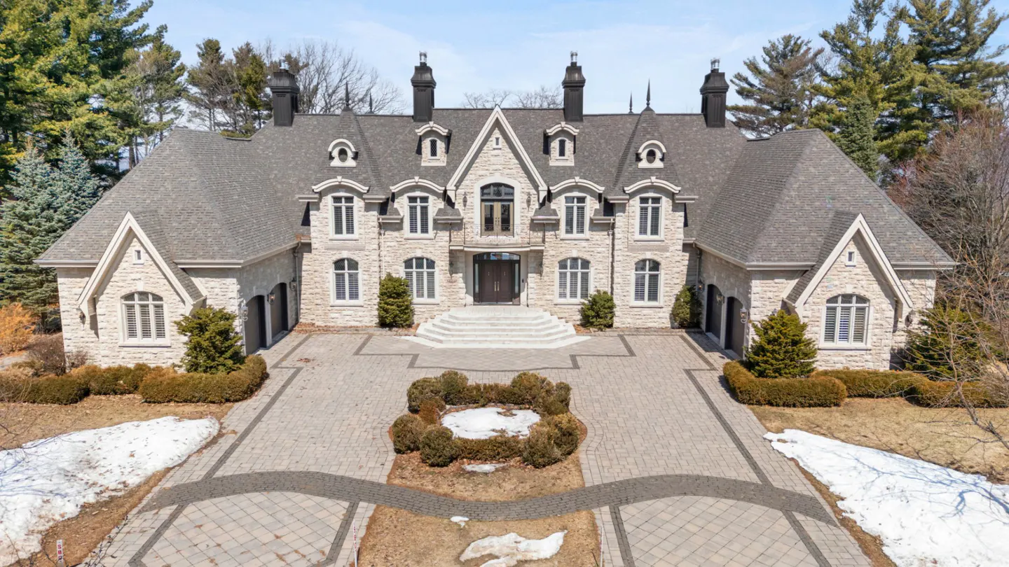 Stone mansion with gray roof, black chimneys, and white-framed windows. Paved driveway with a circular garden in front.