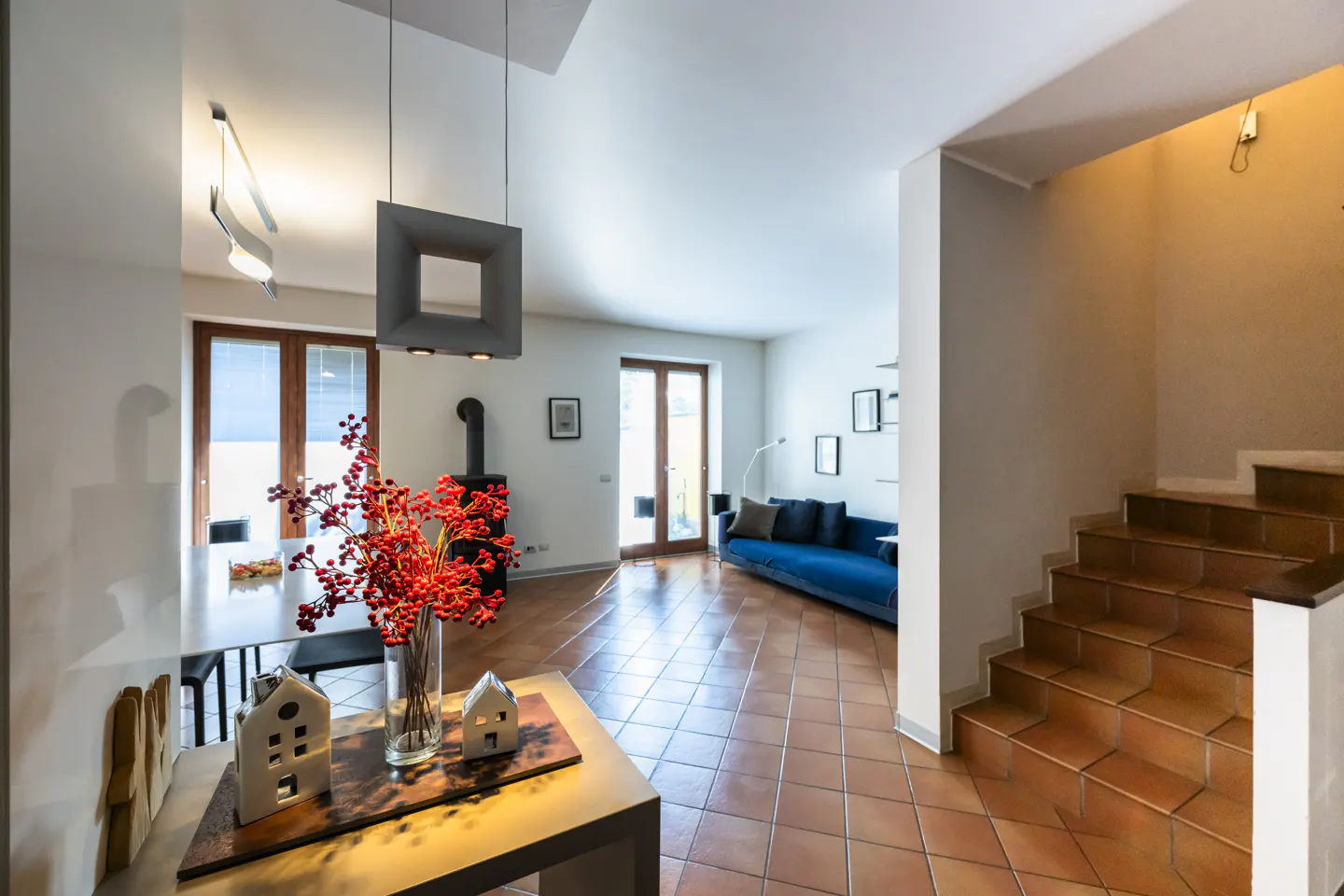 A bright, modern home interior with terracotta tile floors, a blue sofa, and a staircase. A dining table with red berries is in the foreground.