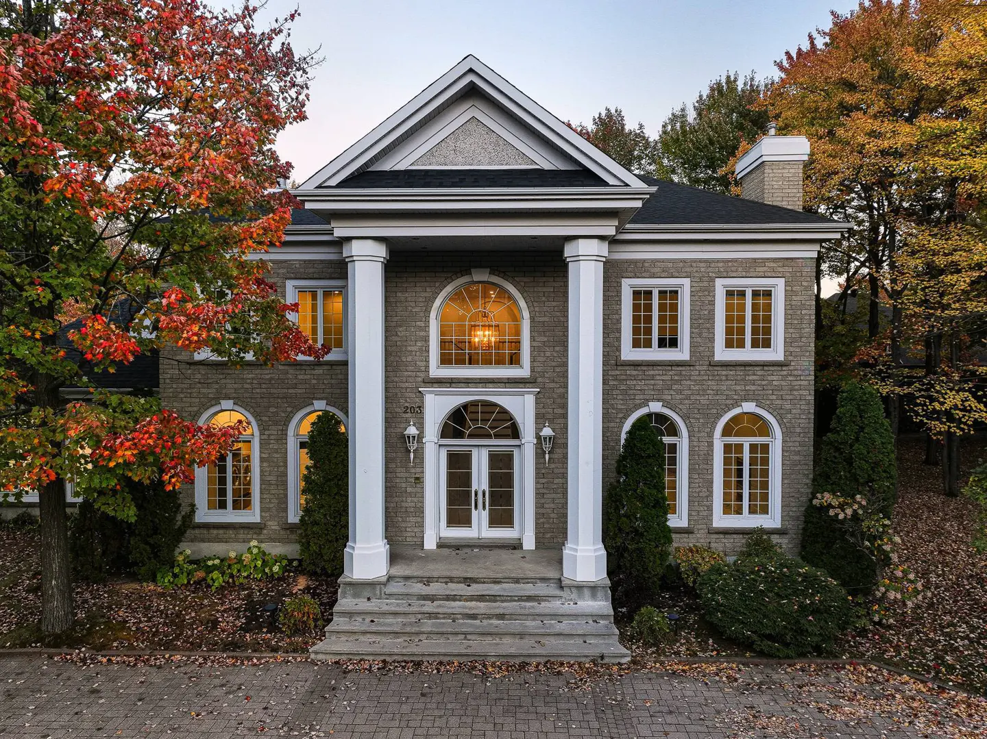 Two-story brick house with white columns and a black roof. Trees with red and orange leaves surround the house.