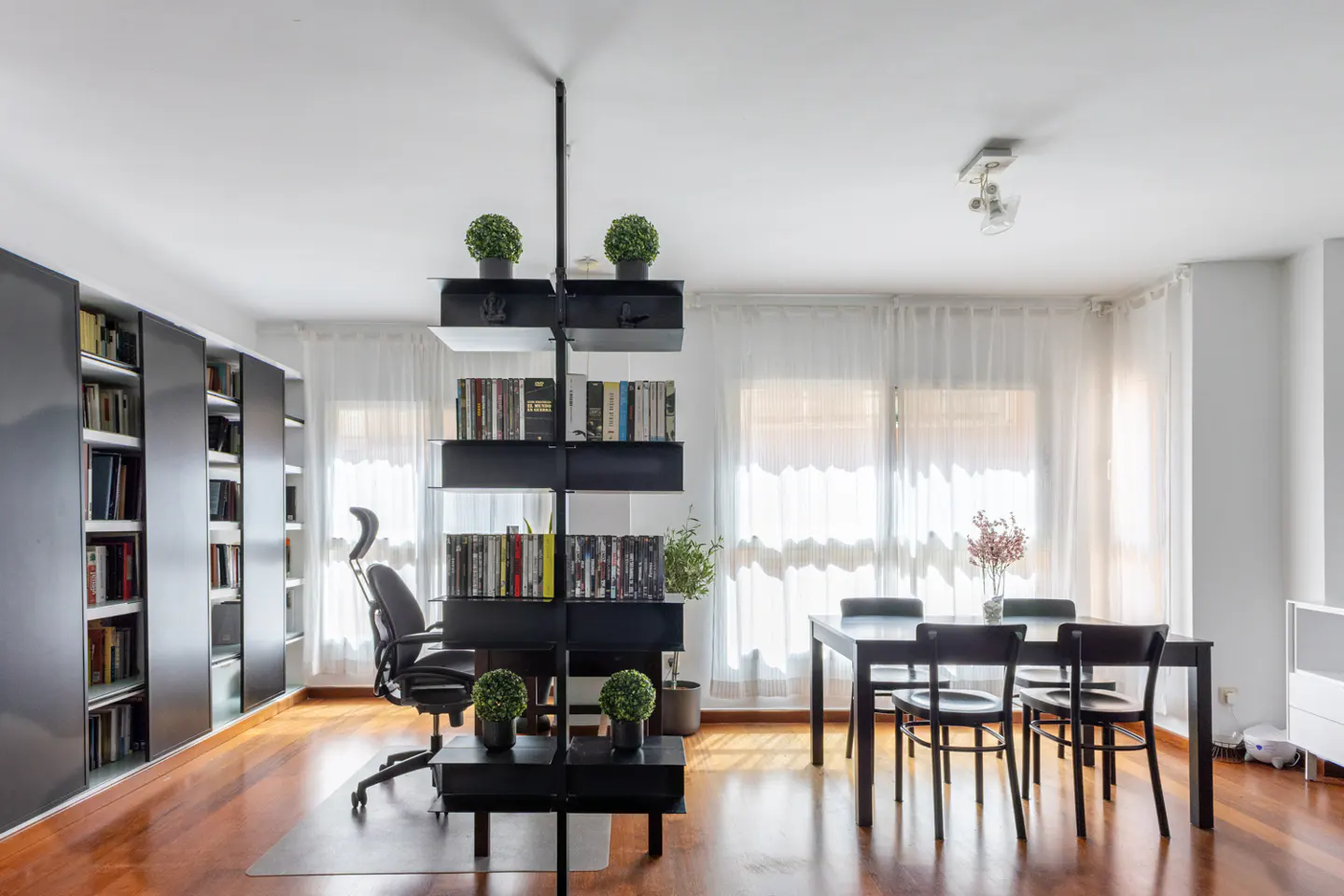 Bright living room with wood floors, black bookshelves, a black dining table with chairs, and white walls.