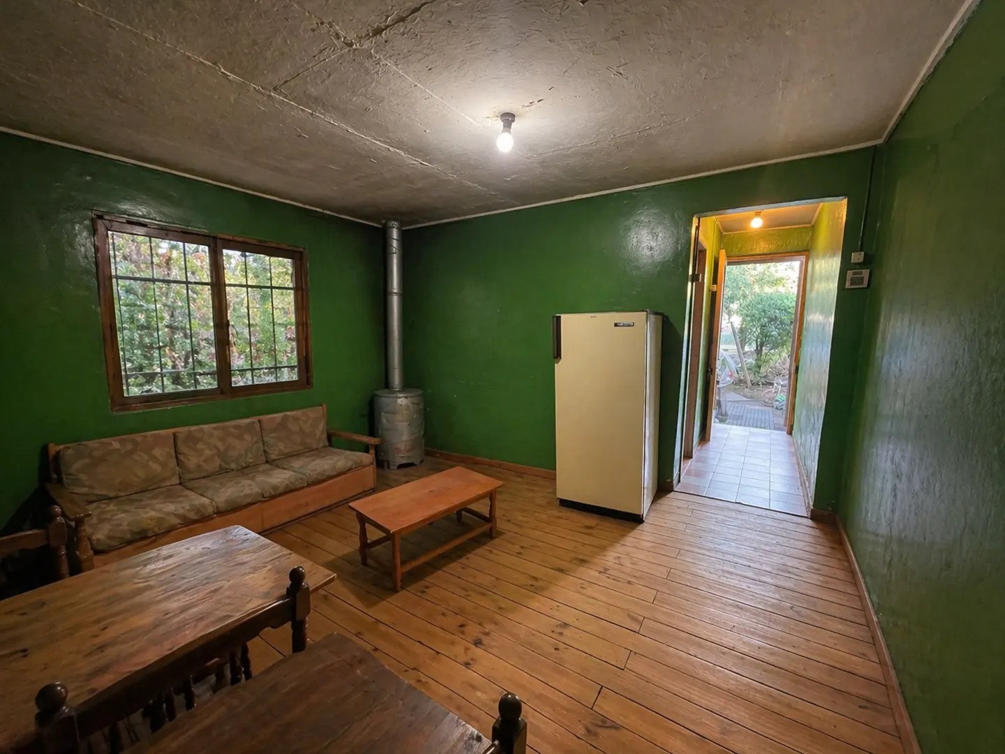A room with green walls, wood floors, and a window. A couch, table, fridge, and wood-burning stove are also present. An open door leads to the outside.