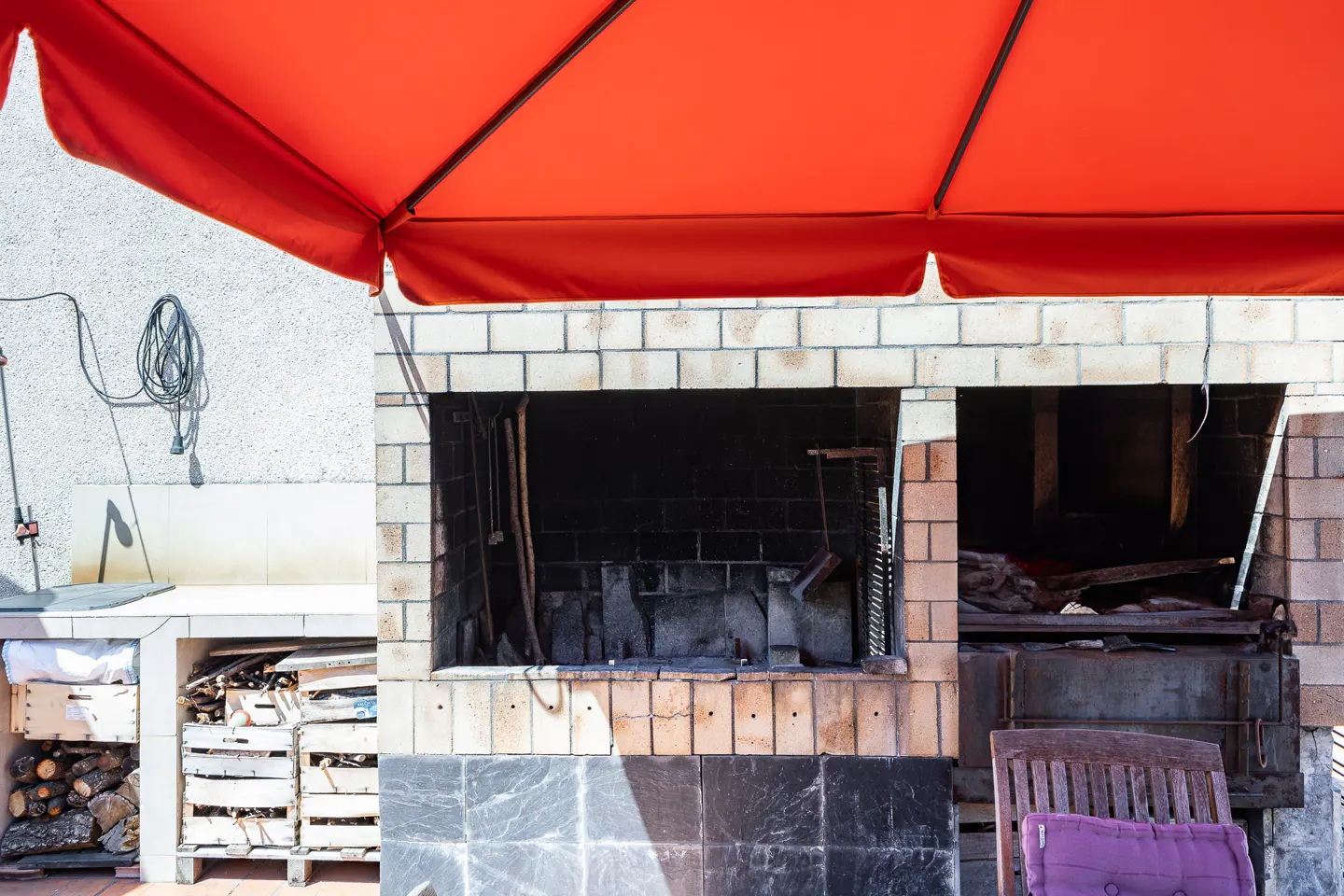Outdoor kitchen with brick grill under a red umbrella. Wood storage and a chair with a purple cushion are visible.