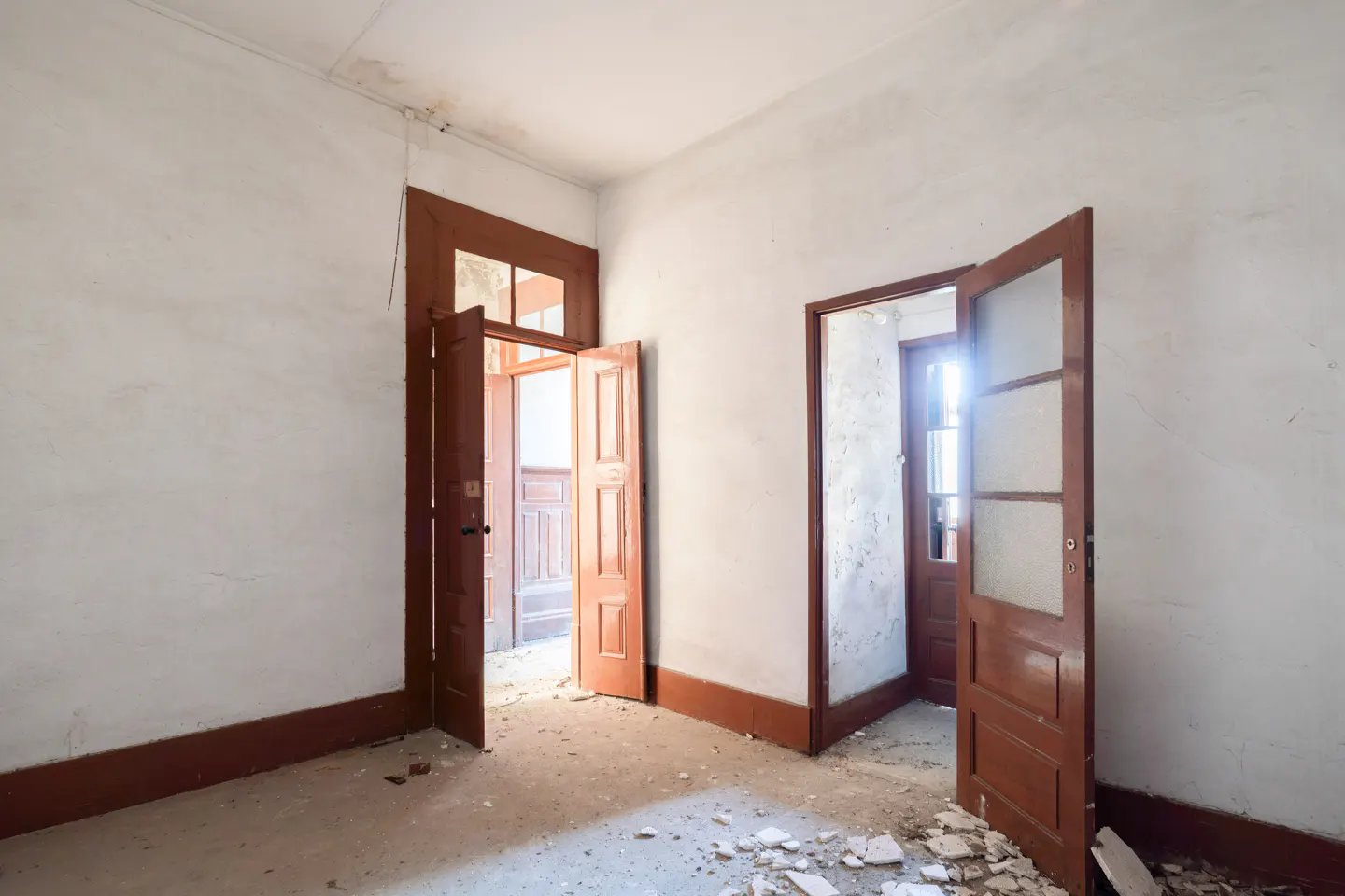 A vacant room with white walls, brown trim, and two open, paneled, brown doors. Debris is scattered on the floor.