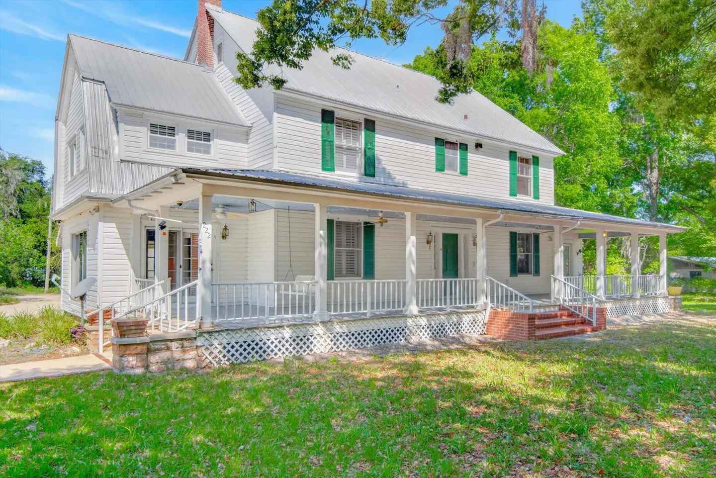 Two-story white house with a metal roof, green shutters, and a long porch with white railings. The lawn is green and trees are in the background.