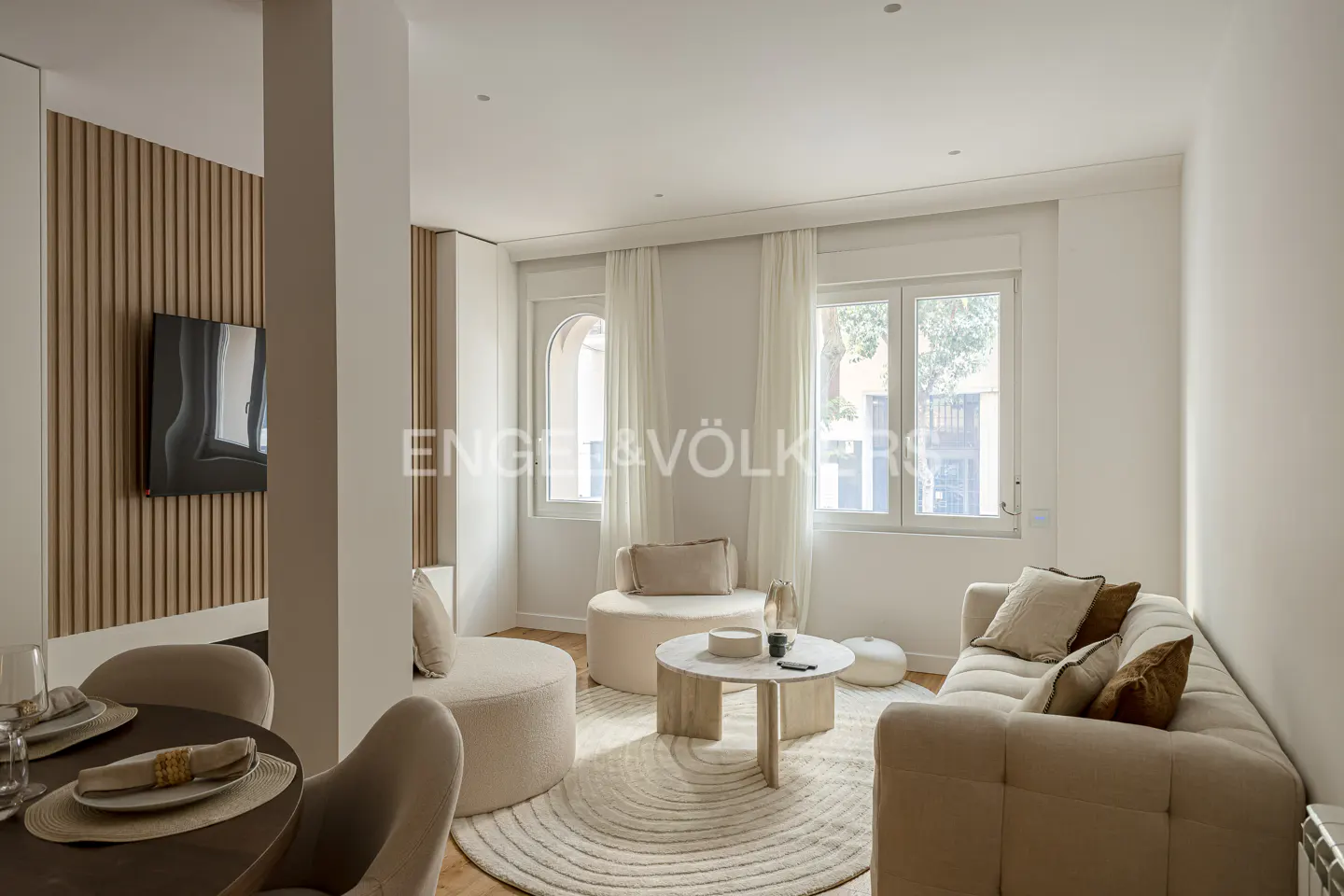 Modern living room with beige sofa, round coffee table, textured wood wall paneling, and white curtains by the windows.