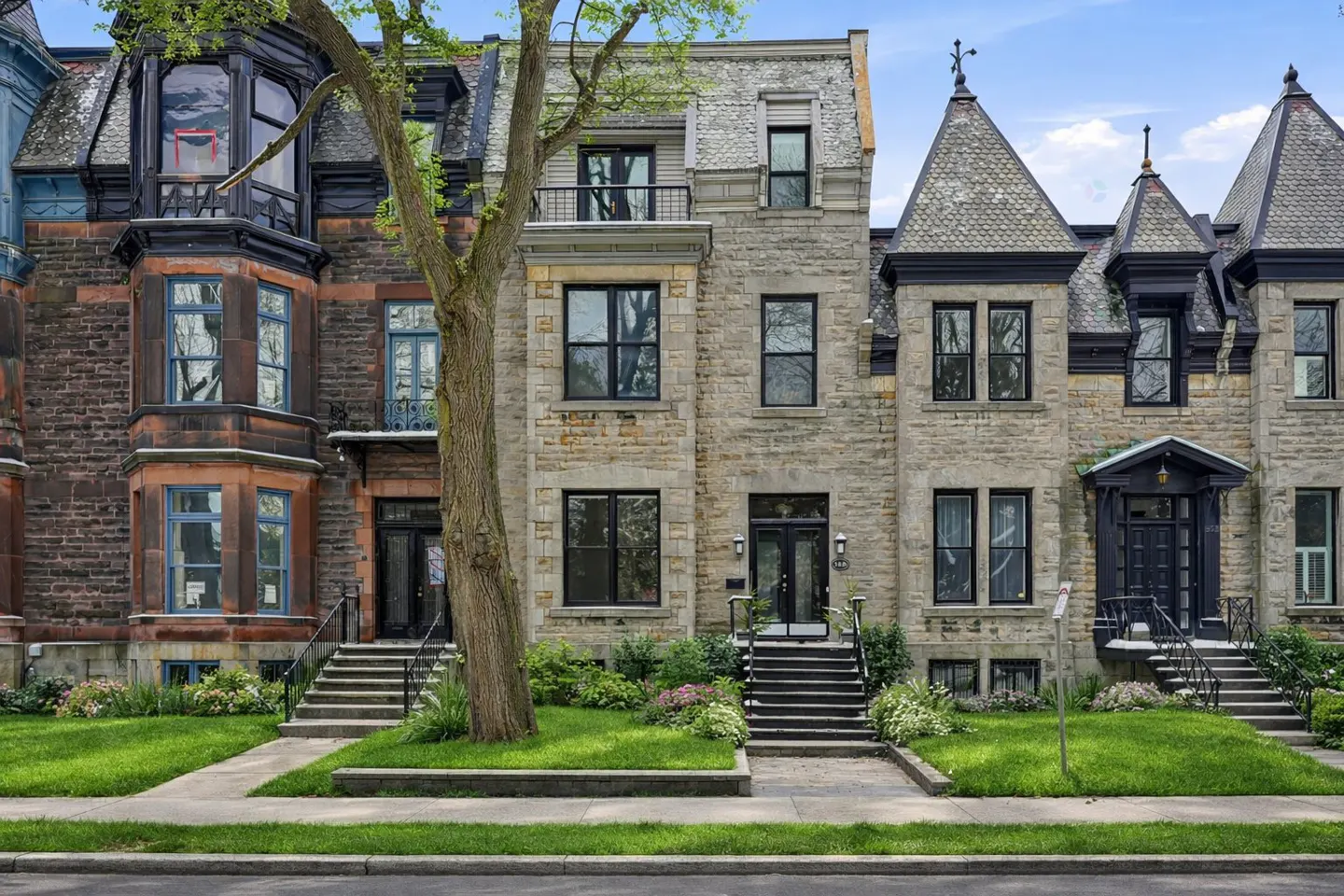 Row of three-story townhouses with stone facades, black doors, and green lawns on a sunny day.