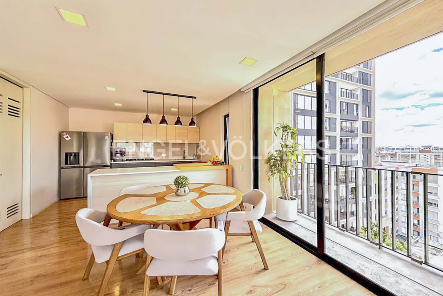 Bright apartment interior with wood floors, a round table with white chairs, and a kitchen with stainless steel appliances. Balcony with city view.