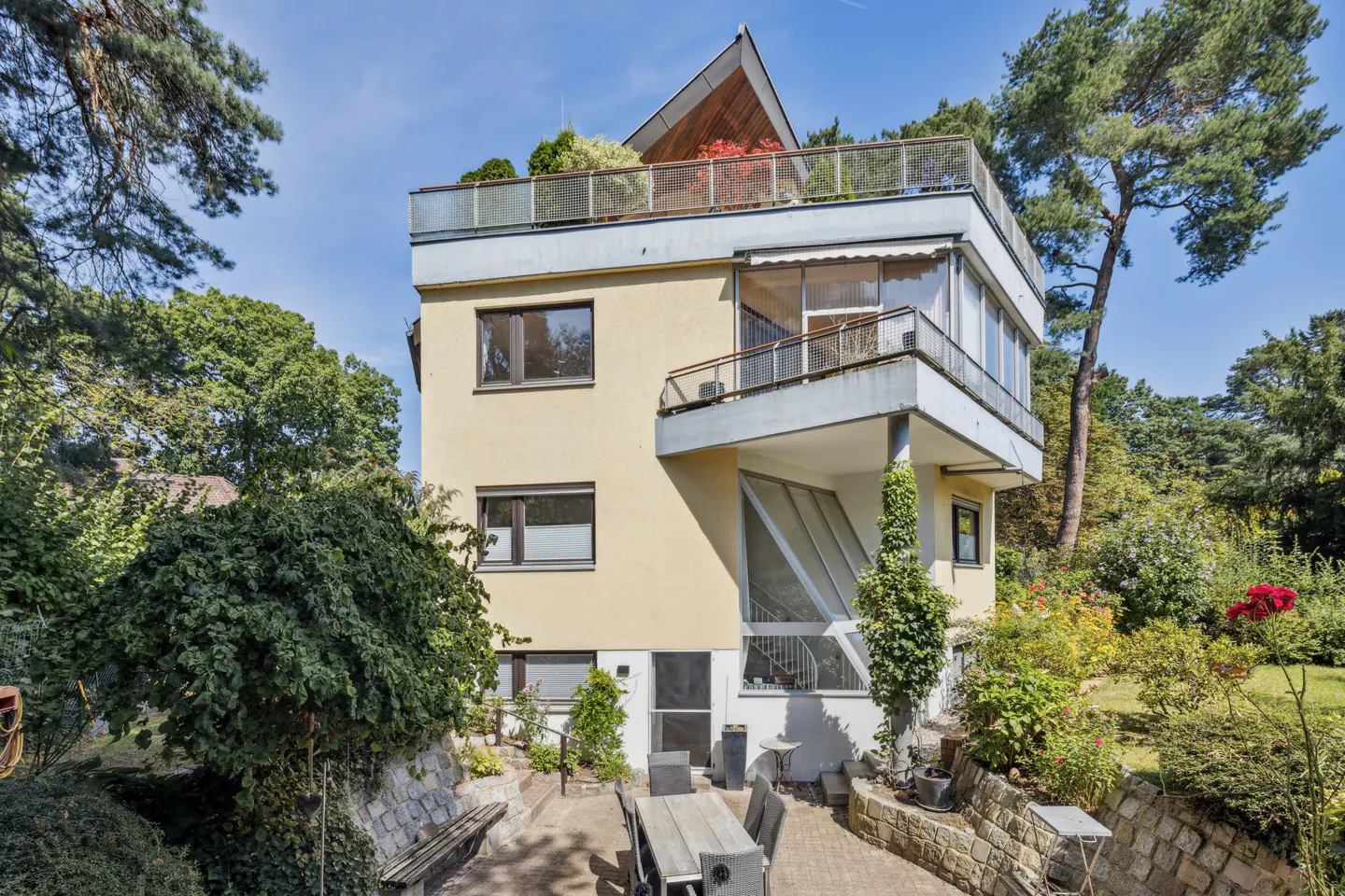 Exterior of a three-story yellow house with a rooftop garden, surrounded by trees and a patio with a table.