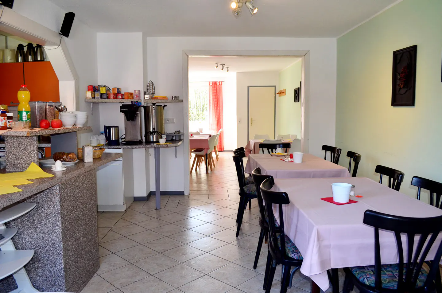 Dining area with tables covered in pink cloths, black chairs, and a breakfast counter with appliances in the background.
