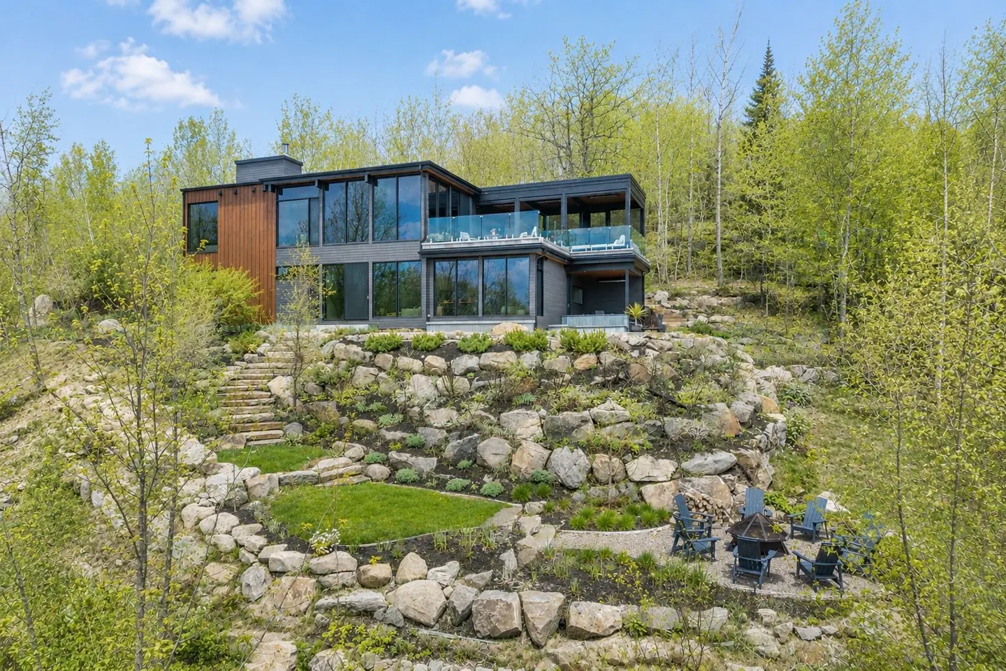 Modern two-story home with wood and dark siding, set on a terraced rocky hillside with a fire pit area and lush trees.